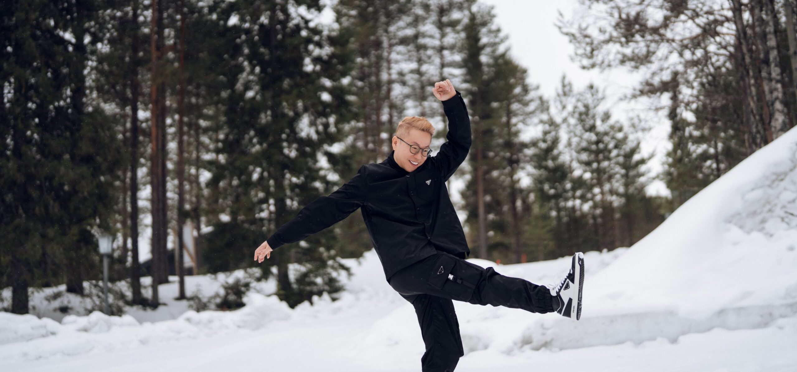 Dan in black, one leg kicked high and a fist in the air, in a snowy pine forest in Finland